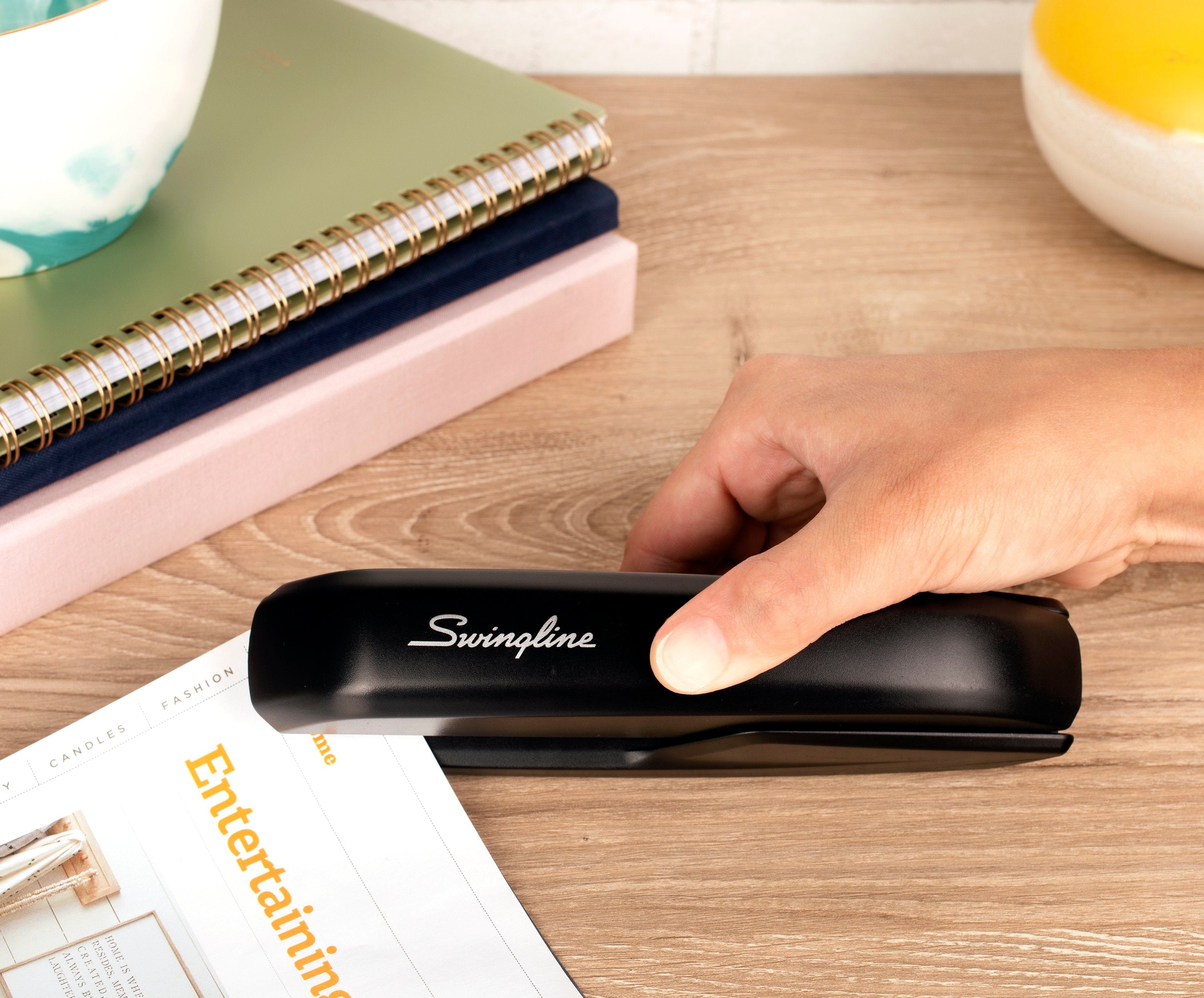 Hand using a Swingline stapler on a desk with stationery items.